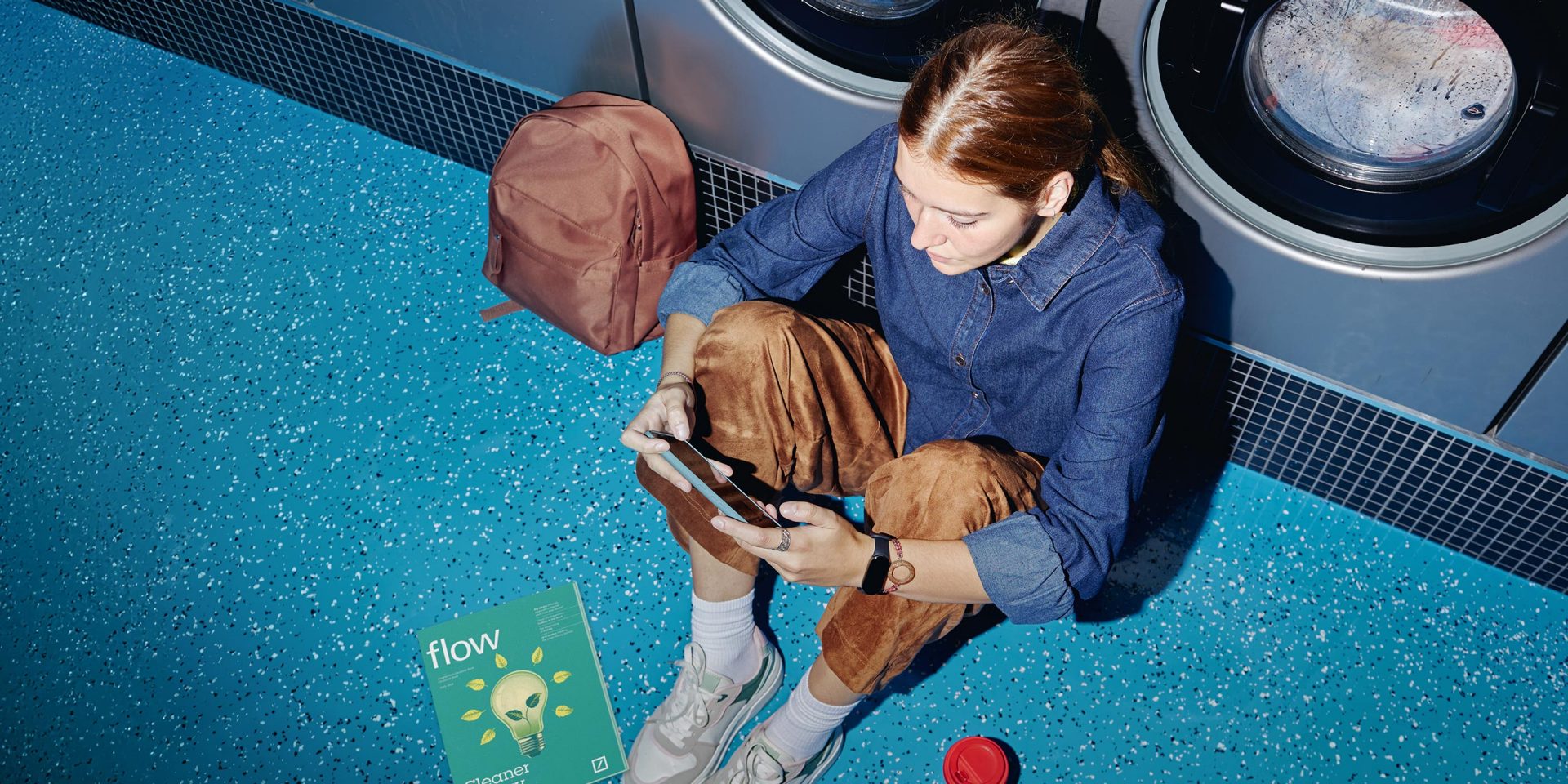 Junge Frau sitzt auf dem Boden eines Waschsalons und nutzt ihr Smartphone um einen Festgeld-Vergleich durchzuführen.