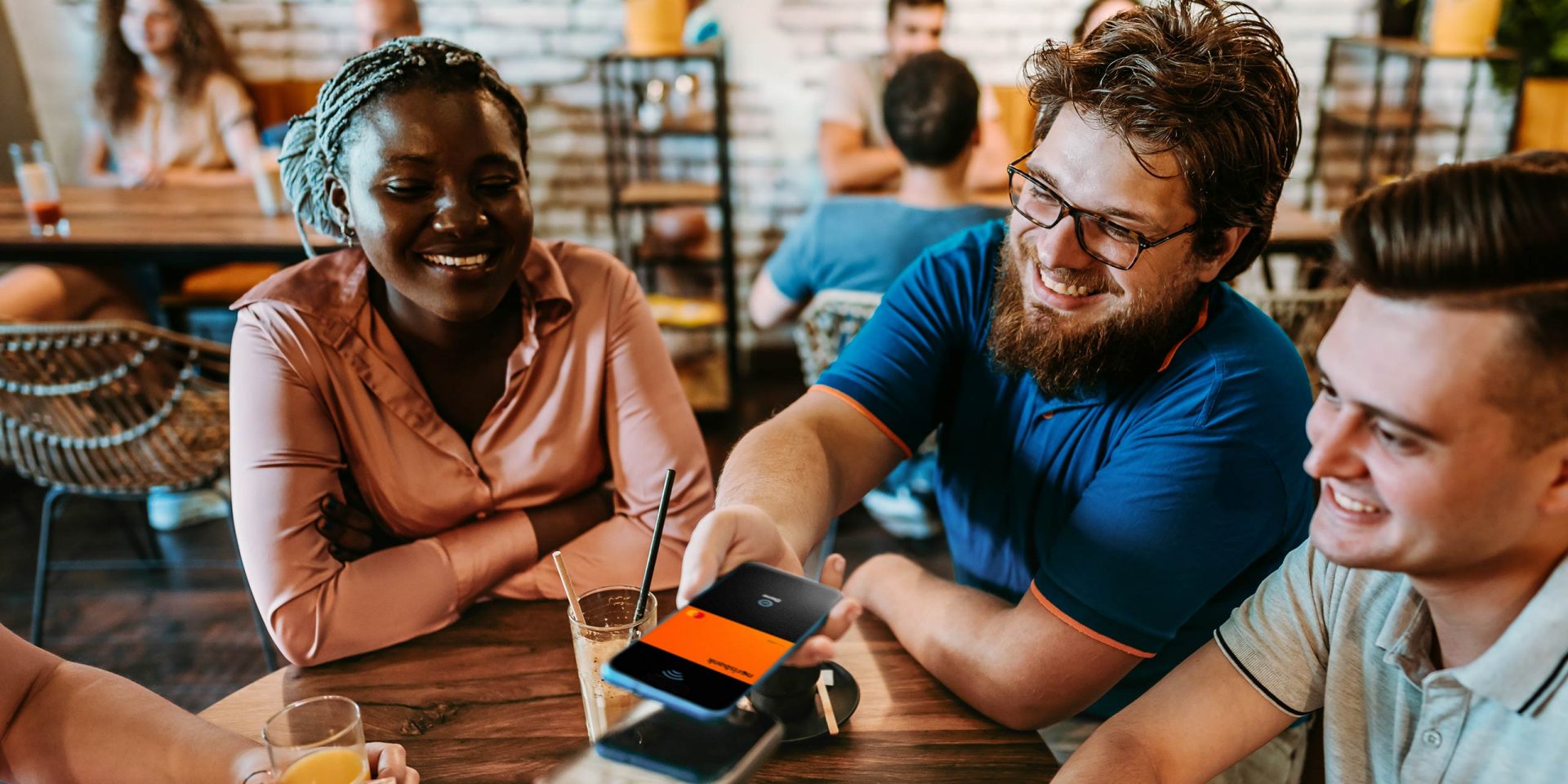 Eine gemischte Gruppe junger Menschen sitzt in einem Restaurant und ein junger Mann hält sein Smartphone an ein Lesegerät, um einen Bezahlvorgang mit Google Pay durchzuführen.