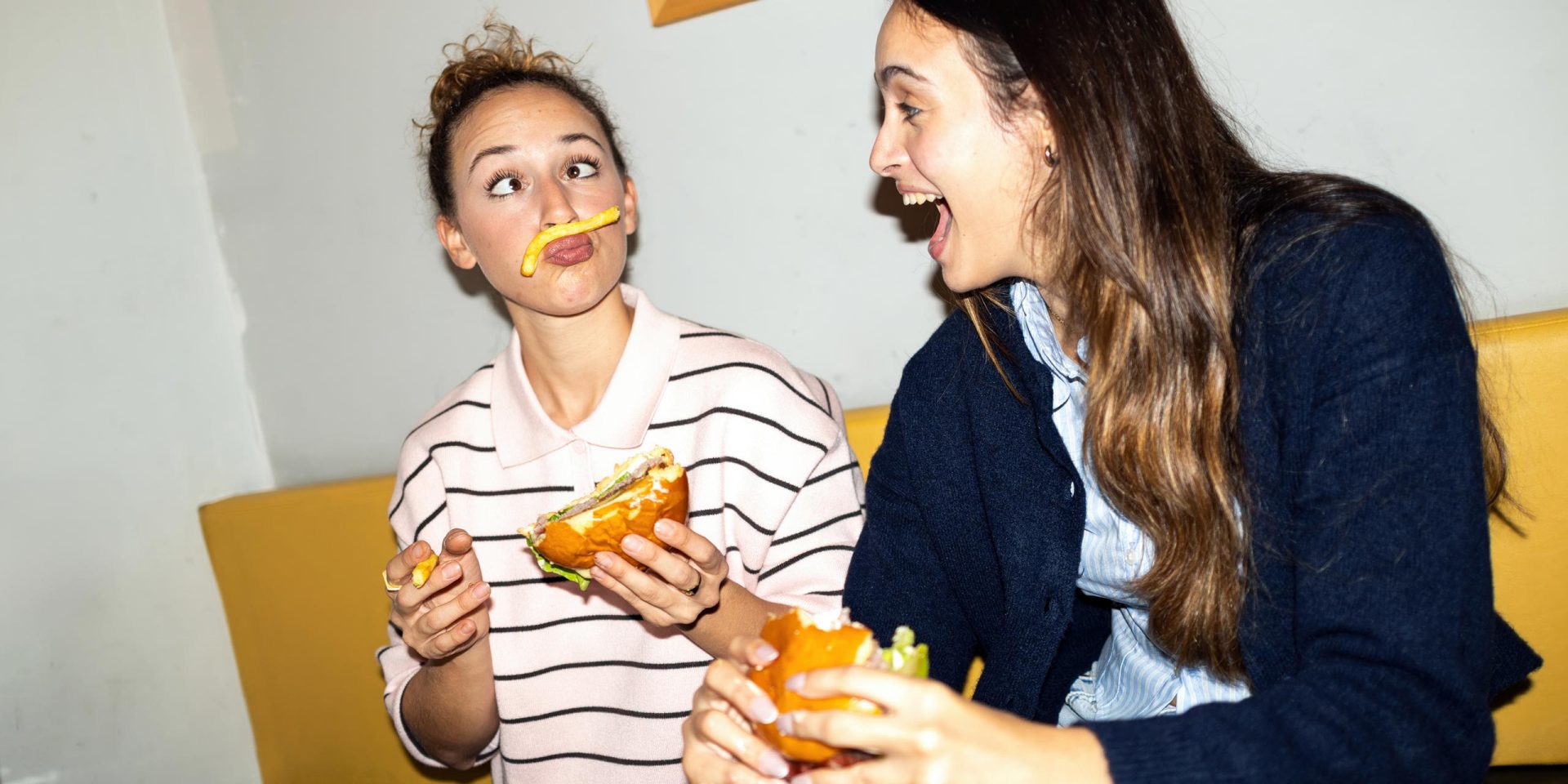 Symbolbild für "kostenloses Girokonto": Zwei Freundinnen in einem Fast Food Restaurant, die sich spielerisch einen Pommes-Schnurrbart  machen, der für Gelächter sorgt.