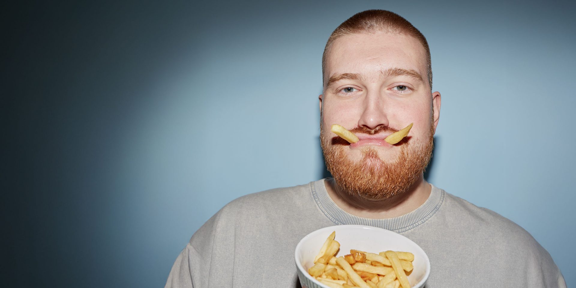 Symbolbild für "Ratenkredit": Porträt eines jungen erwachsenen Mannes, der mit zwei Pommes im Mund ein lustiges Gesicht macht.