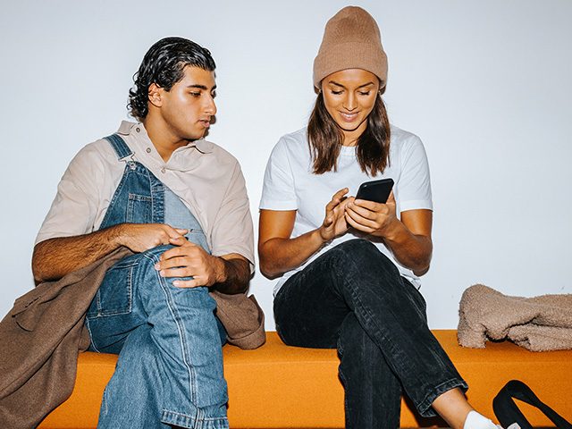 Zwei Studenten sitzen auf einer Bank und die Studentin zeigt auf ihrem Smartphone ihr Studentenkonto.