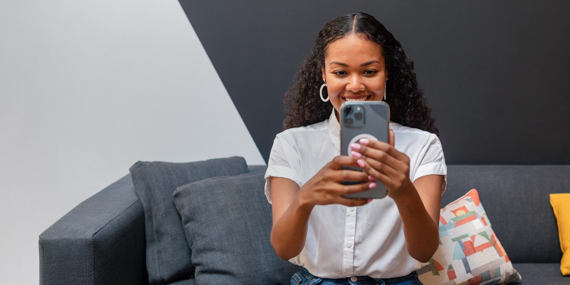 Eine junge Frau sitzt auf ihrem Sofa und lächelt in die Kamera ihres Smartphones. Sie durchläuft den Prozess einer Video-Legitimation zum Abschluss eines norisbank-Produkts.