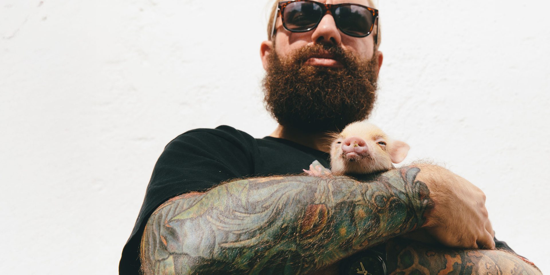 Mann mit tätowierten Armen und Sonnenbrille hat ein kleines Ferkel auf dem Arm, symbolhaft für "Weltspartag".