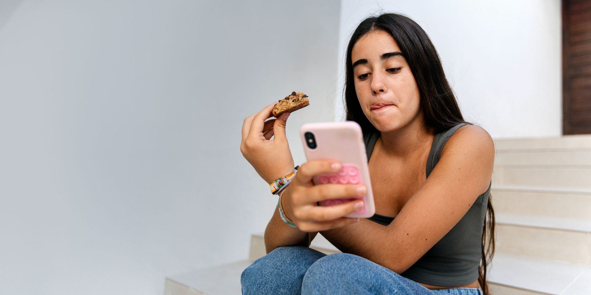 Junge Frau sitzt entspannt, isst einen Cookie und nutzt ein rosa Smartphone, Alltagsszene mit Fokus auf mobile Nutzung von wero