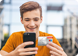 Teenager-Junge hält stolz Handy und eine Maestro Karte (Debitkarte) in den Händen.